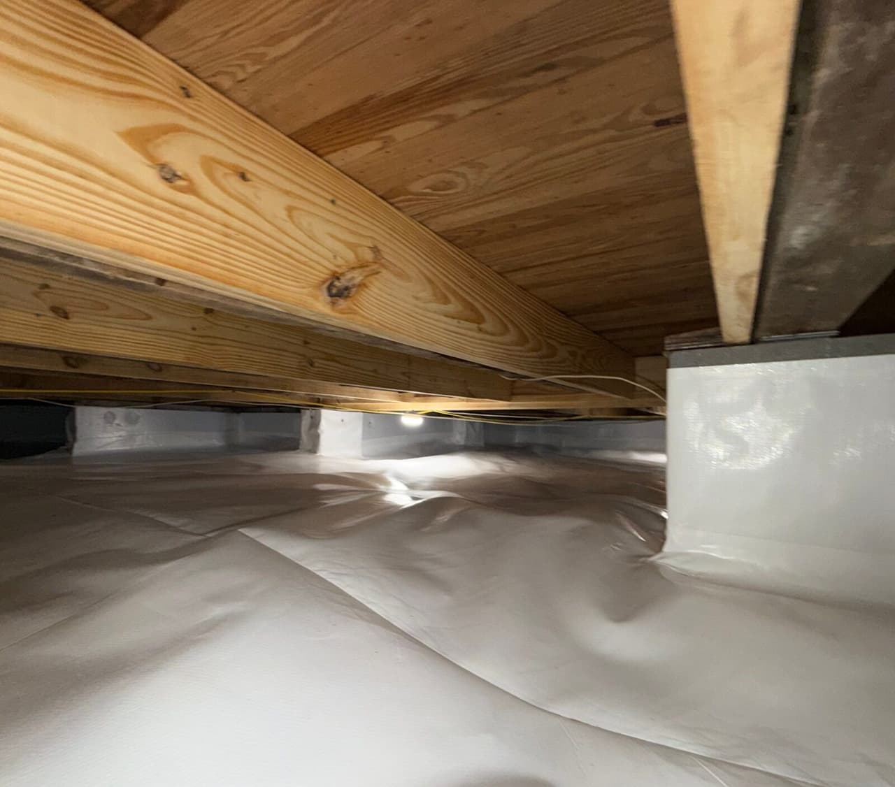 Crawl space with white liner on the floor, a wrapped pier and new lumber joists above.