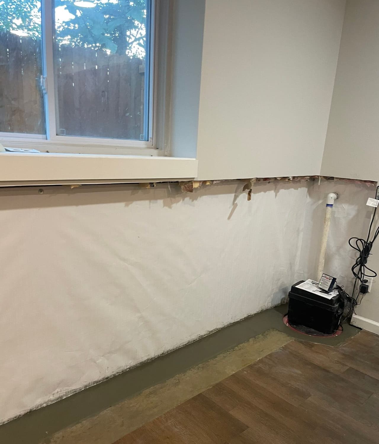 Basement wall with vapor barrier, patched floor drain trench, and a sump pit with lid and battery backup box.
