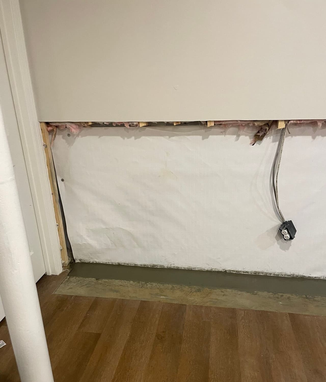 Basement wall section with vapor barrier below cut drywall and a patched drain trench at the floor edge.