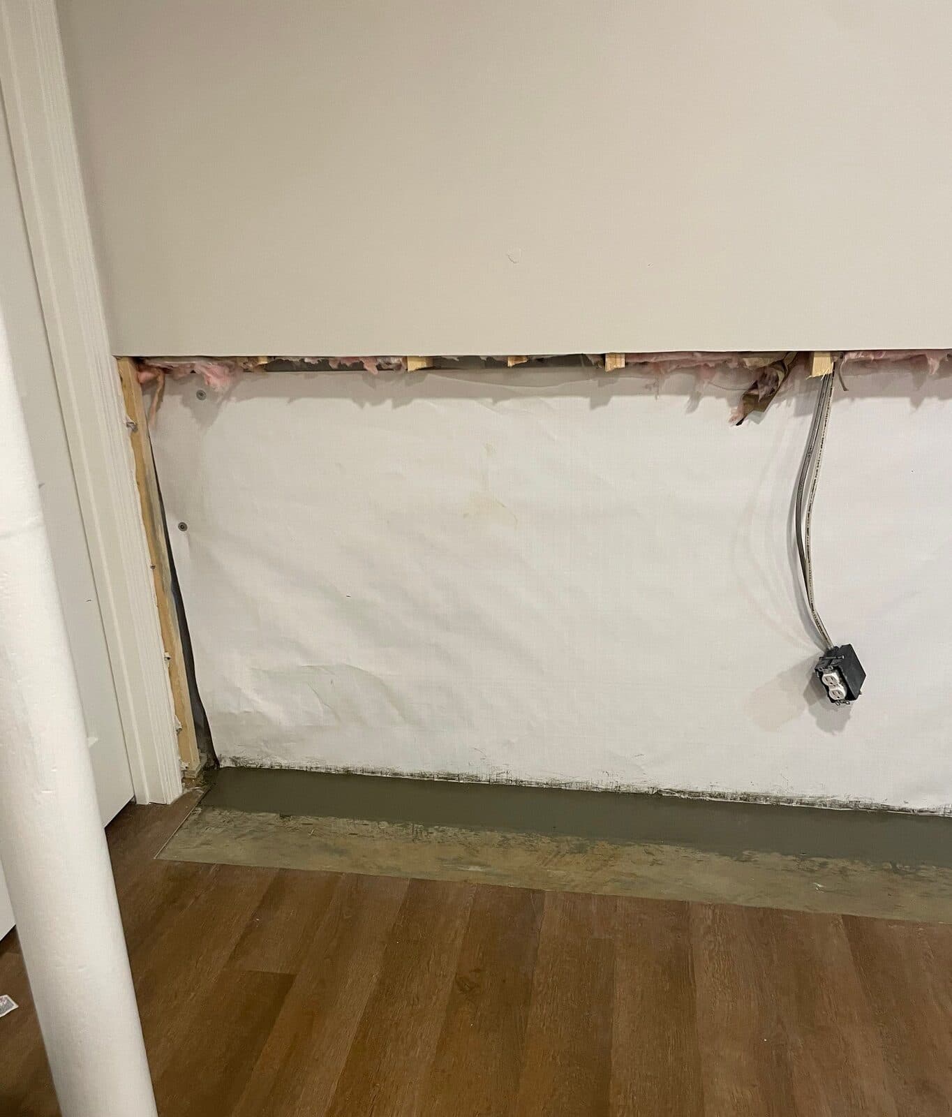 Basement wall section with vapor barrier below cut drywall and a patched drain trench at the floor edge.