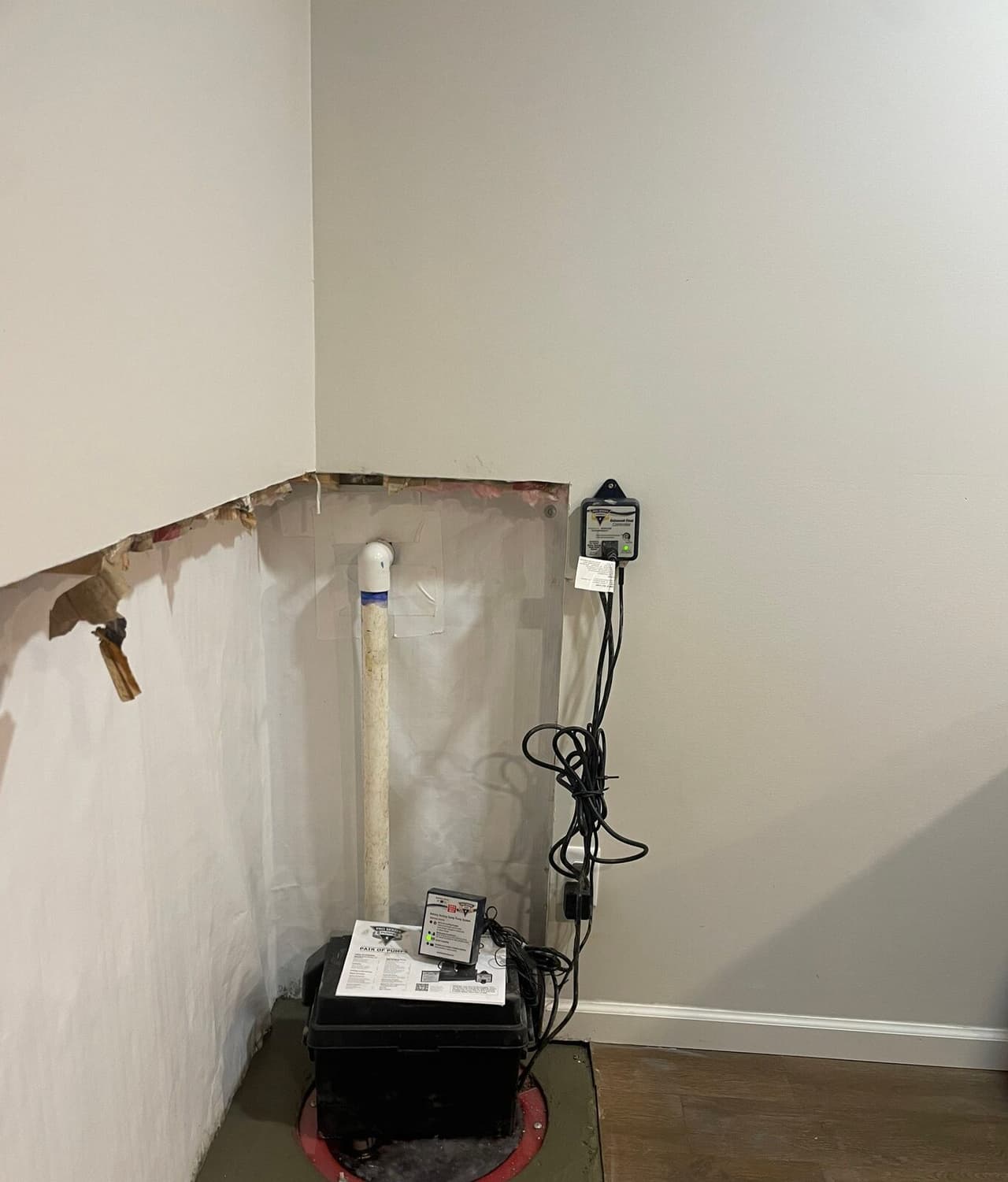 Close view of a sump pump battery backup box on a lidded sump pit with a discharge pipe and controller.