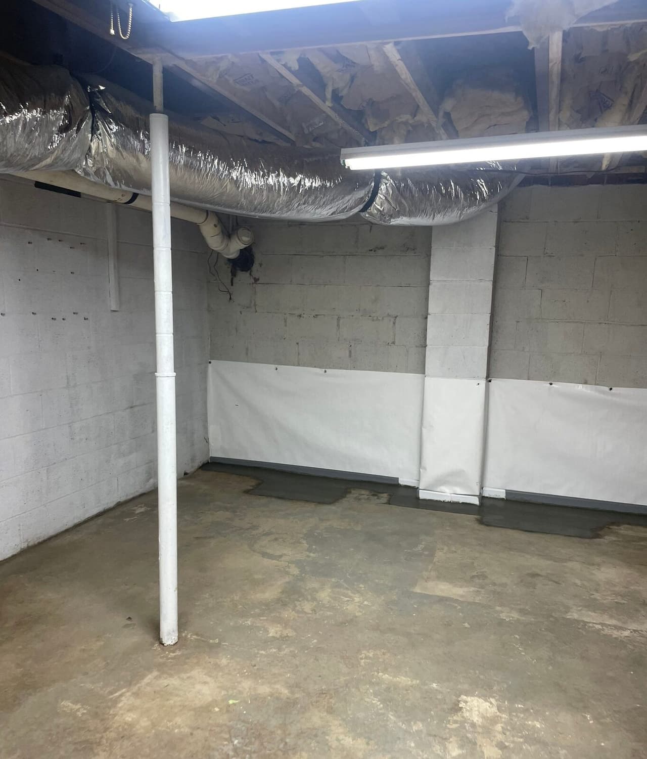 Basement room with white liner along lower walls, patched floor edge, a support post and overhead ductwork.