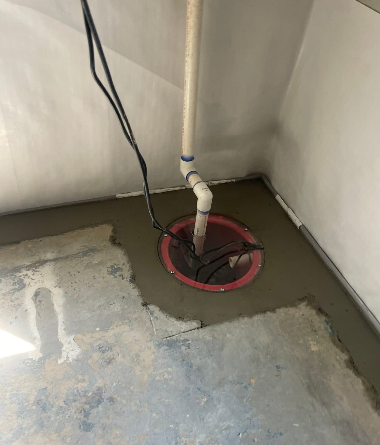 Sump pit with sealed lid and discharge pipe set in fresh concrete in a lined basement corner.