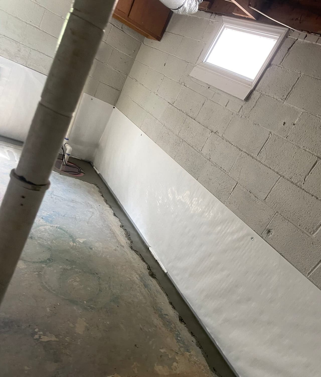 Tilted view of a basement wall with white liner, patched floor edge and a sump in the background.