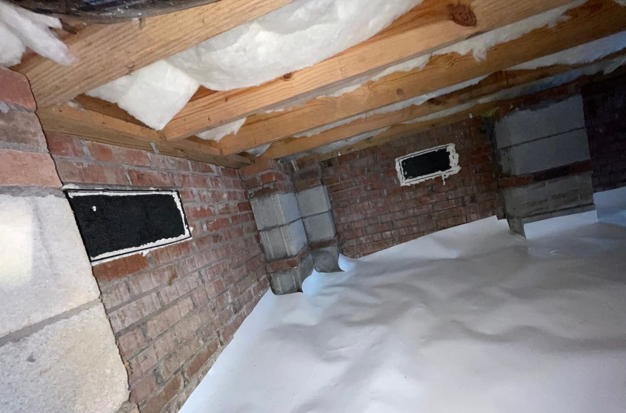 Crawl space corner with white floor liner against brick walls and foam sealed vent openings.