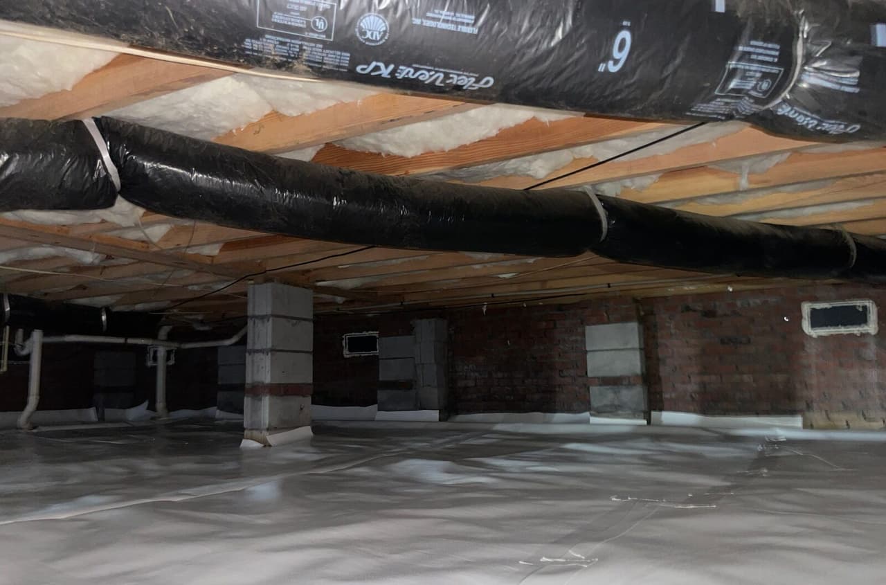 Wide crawl space view with white liner on the floor and piers and ducts running under the joists.