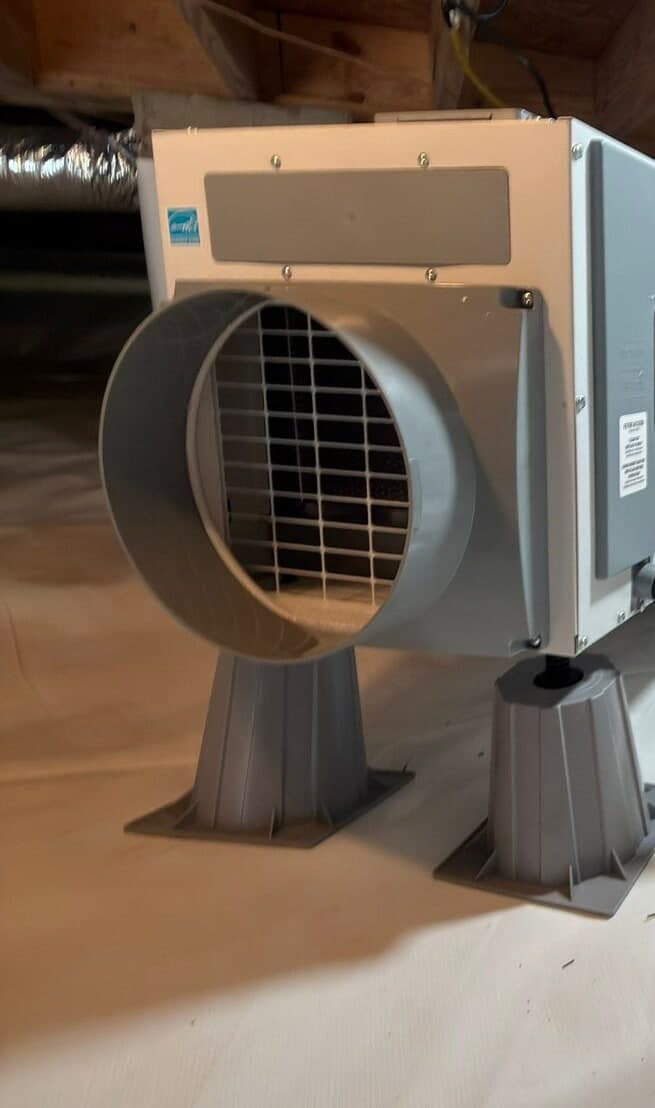 Dehumidifier on gray stands over a white crawl space liner beneath the floor joists.