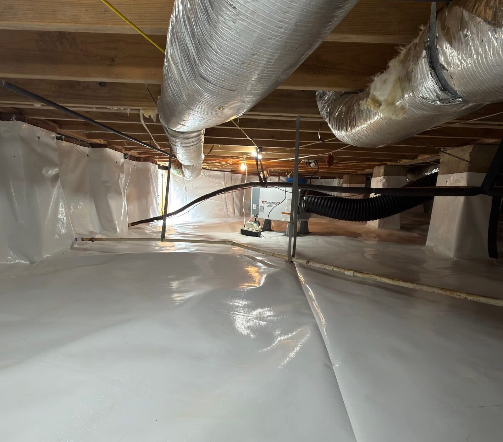 Lined crawl space with a dehumidifier set on the liner beneath ducts and joists.