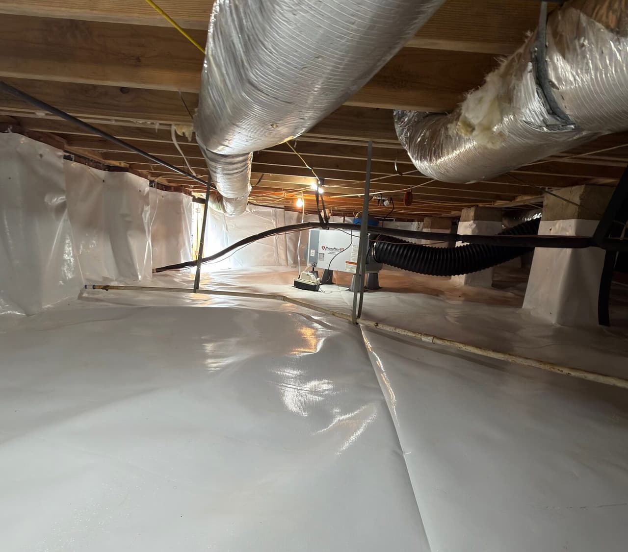 Lined crawl space with wrapped piers and a dehumidifier installed under silver ducts.
