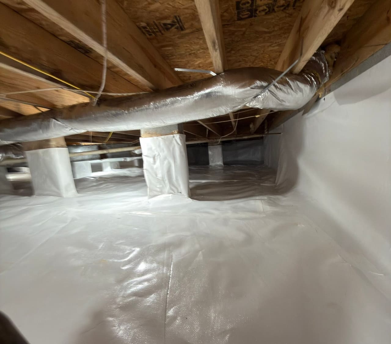 Crawl space with white liner on the floor and walls and wrapped piers under a silver duct.