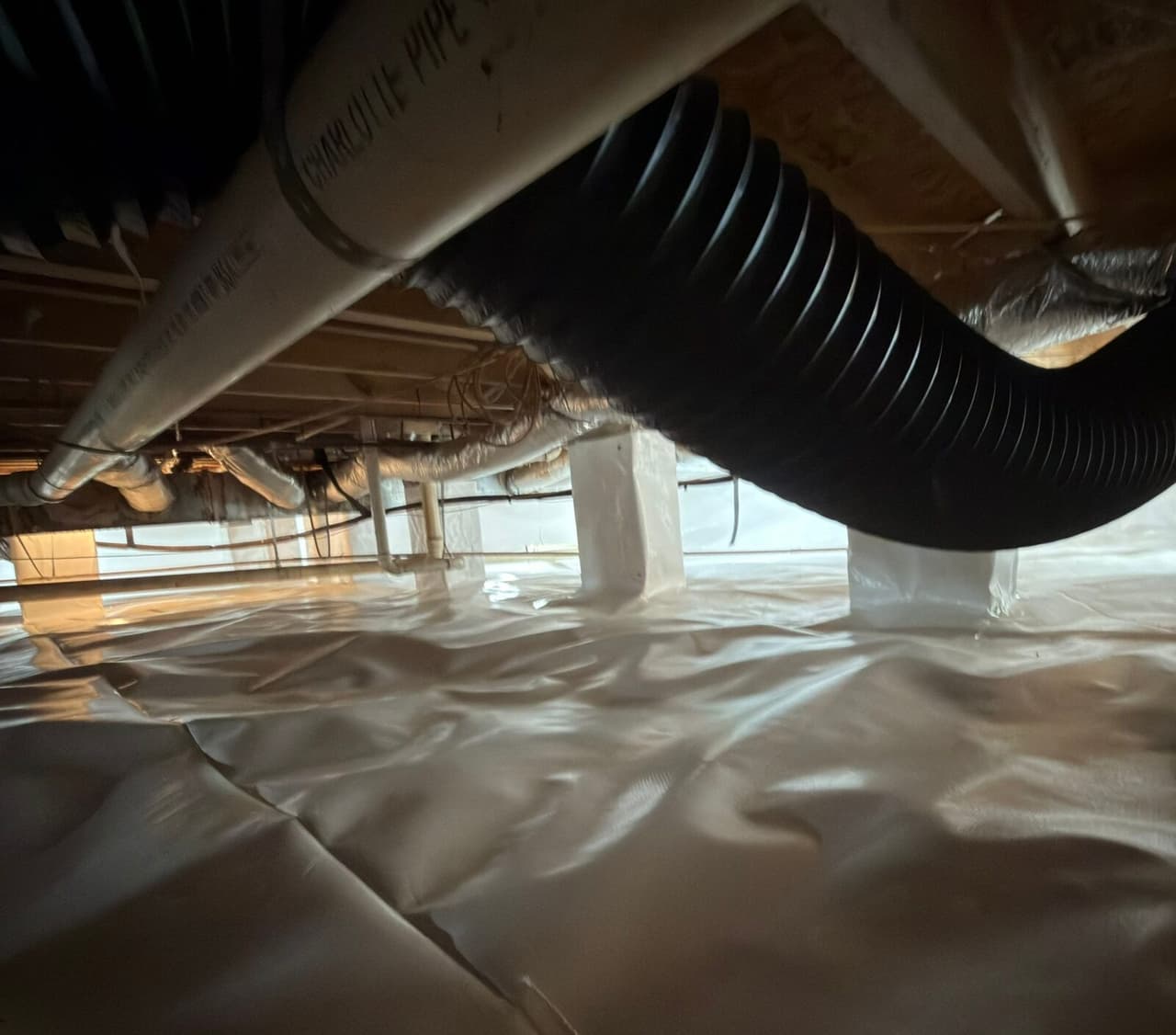 Crawl space with white liner on the floor and wrapped piers under a black corrugated duct and pipes.