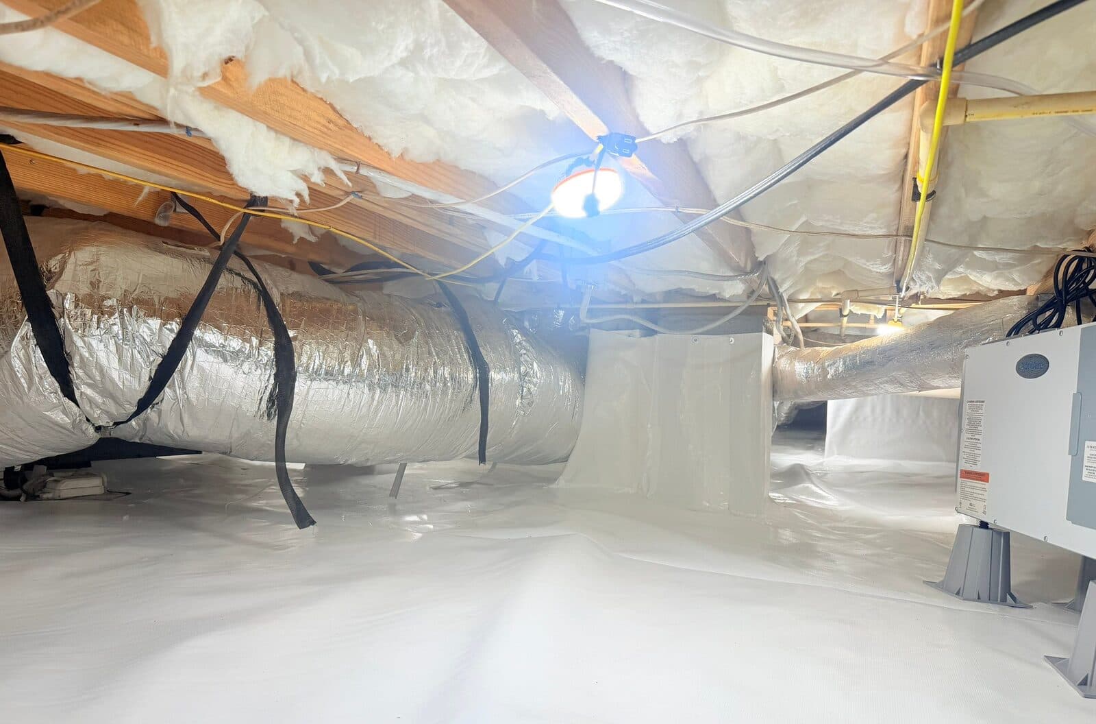 Wide view of a lined crawl space with a foil duct, work light, and dehumidifier at the right edge.