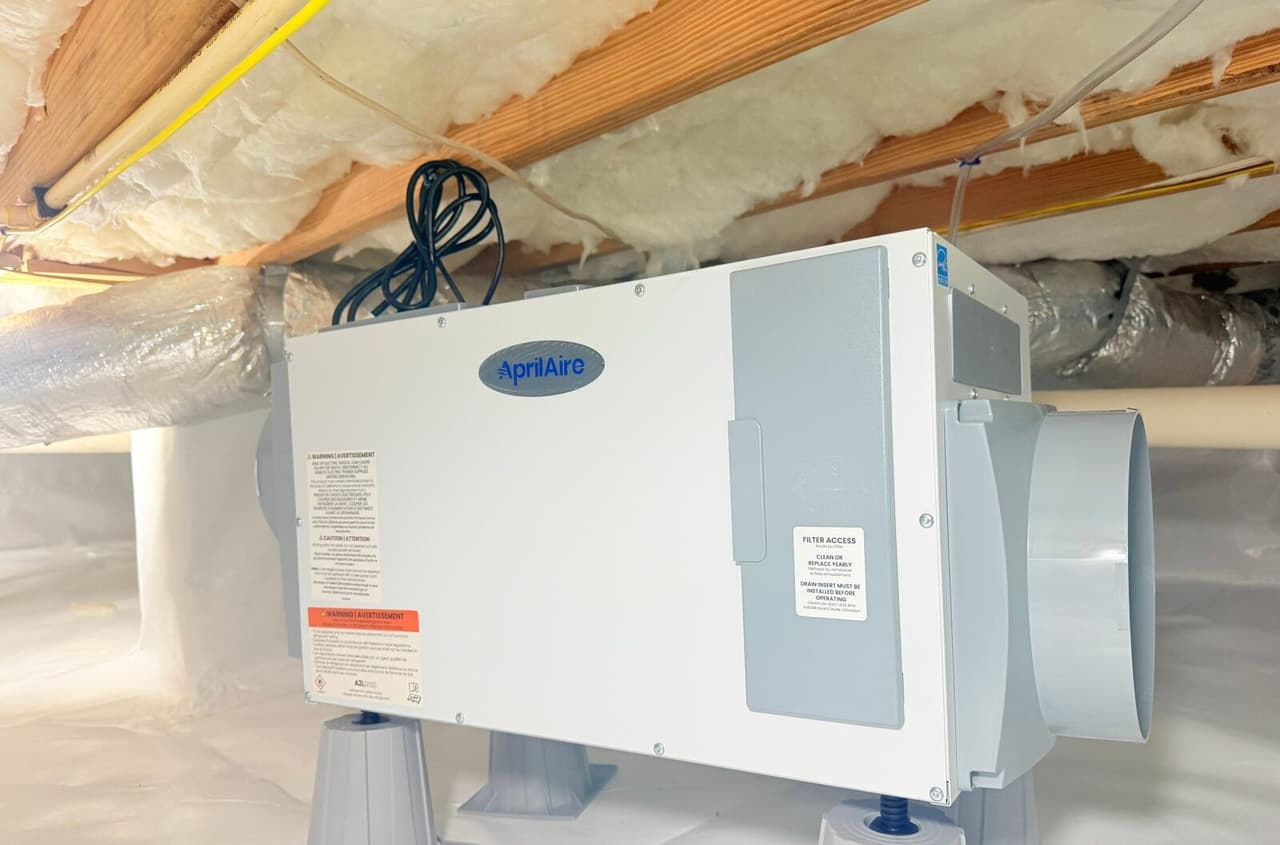 Crawl space dehumidifier mounted on plastic stands over white liner beneath insulated floor joists.