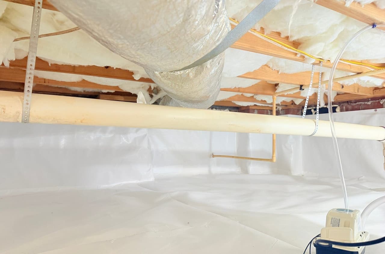 Crawl space with white liner covering the floor and walls, insulated joists above and a condensate pump in the corner.