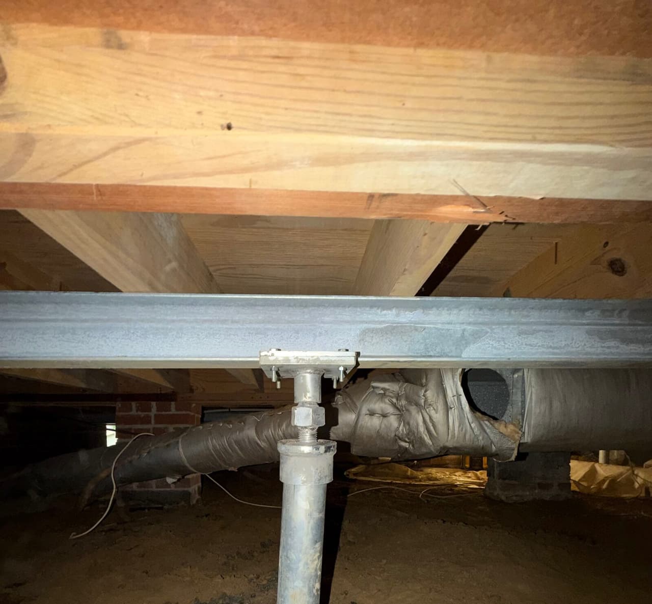 Adjustable steel post bolted to a steel beam beneath a wood girder and floor joists in a crawl space.
