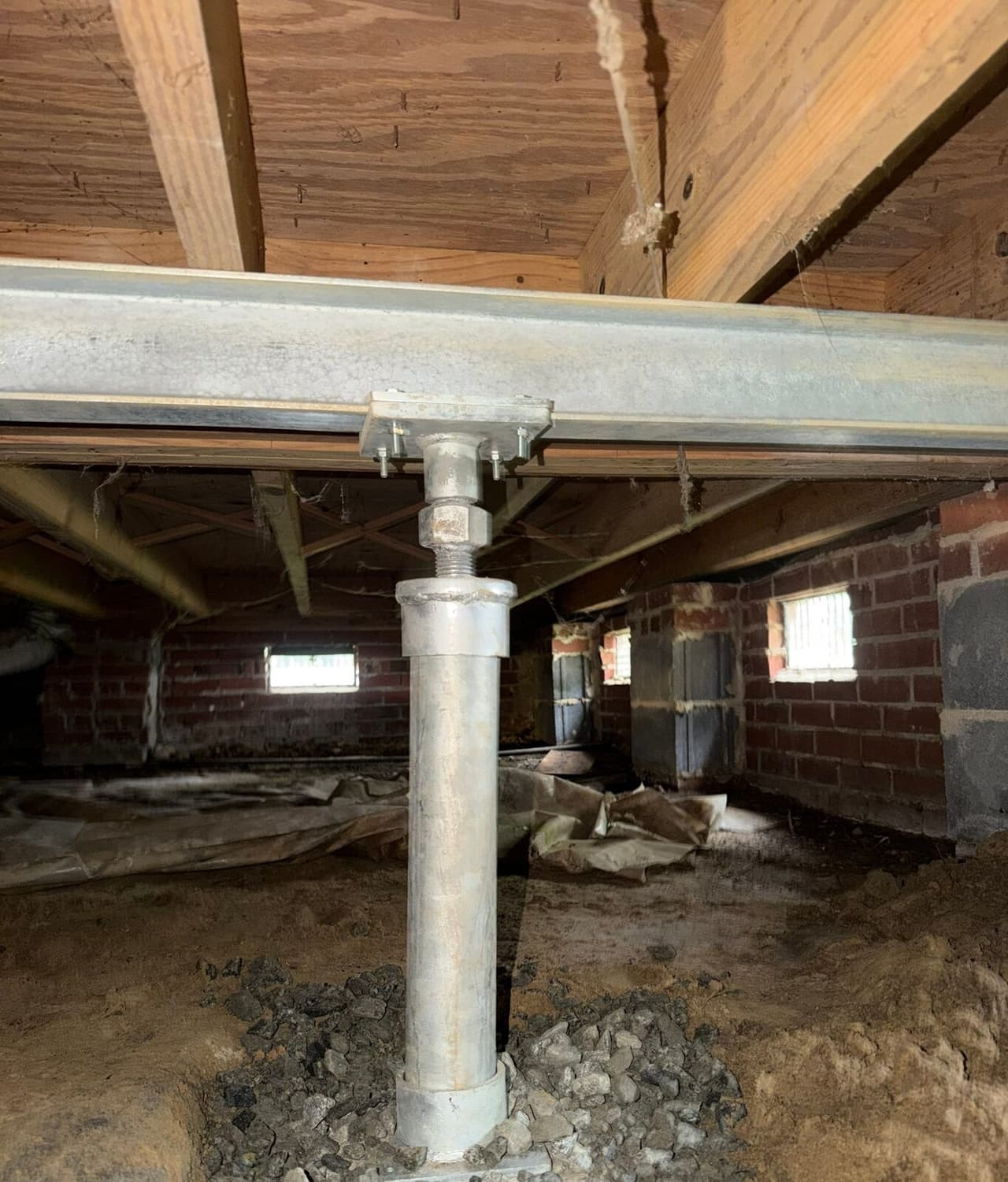 Adjustable steel post on a gravel base bolted to a steel beam under floor joists in a crawl space.