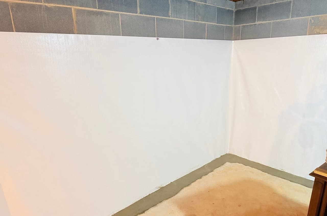 Basement corner with white wall membrane and a patched concrete perimeter strip along the floor.