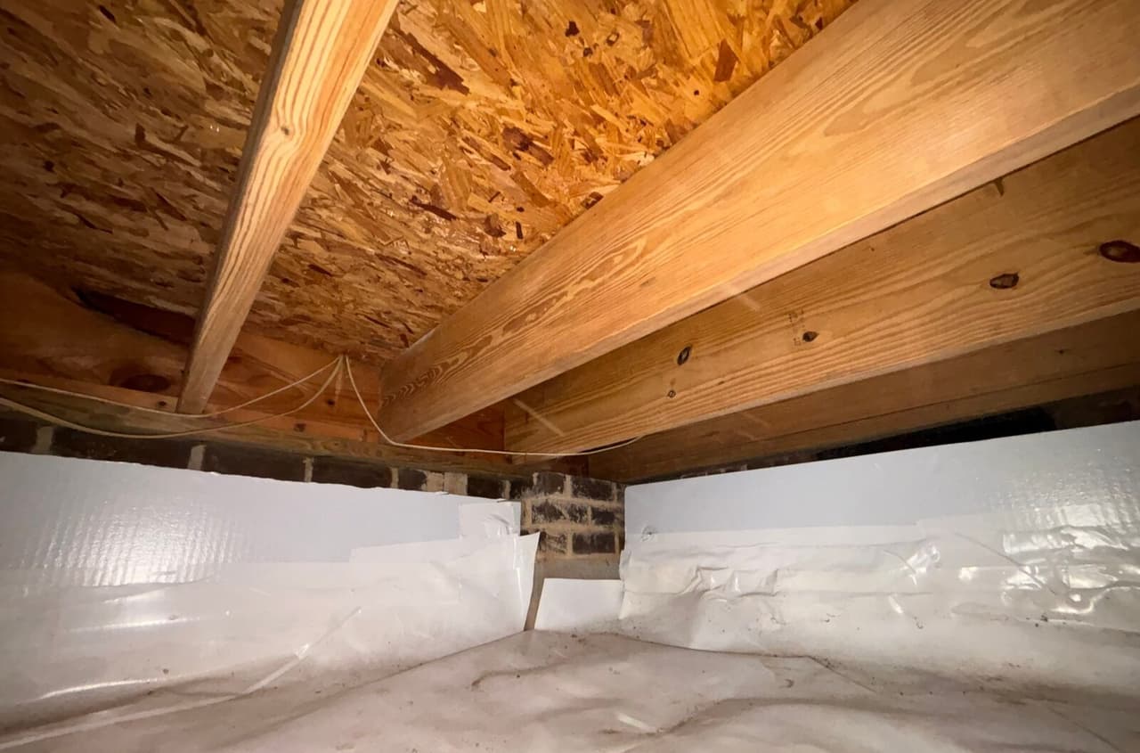Crawl space corner with foam board on walls and white liner covering the floor under joists.
