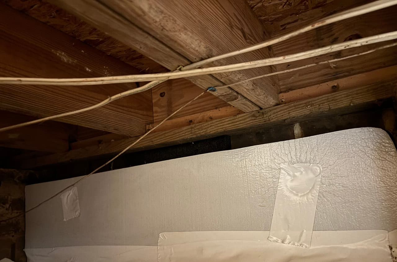 Foam board panel with taped patches along a crawl space wall below wooden joists and wiring.