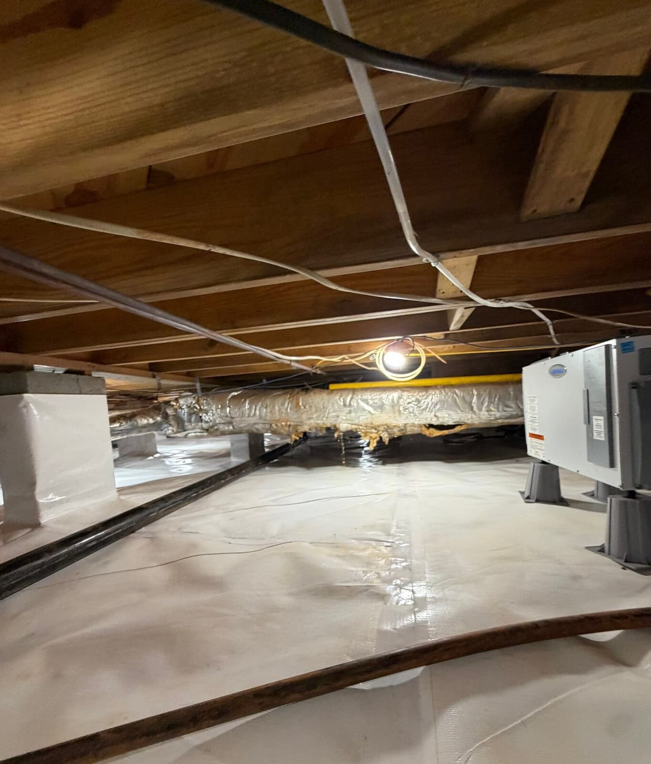 Crawl space with white liner on the floor, wrapped piers and a dehumidifier at right.