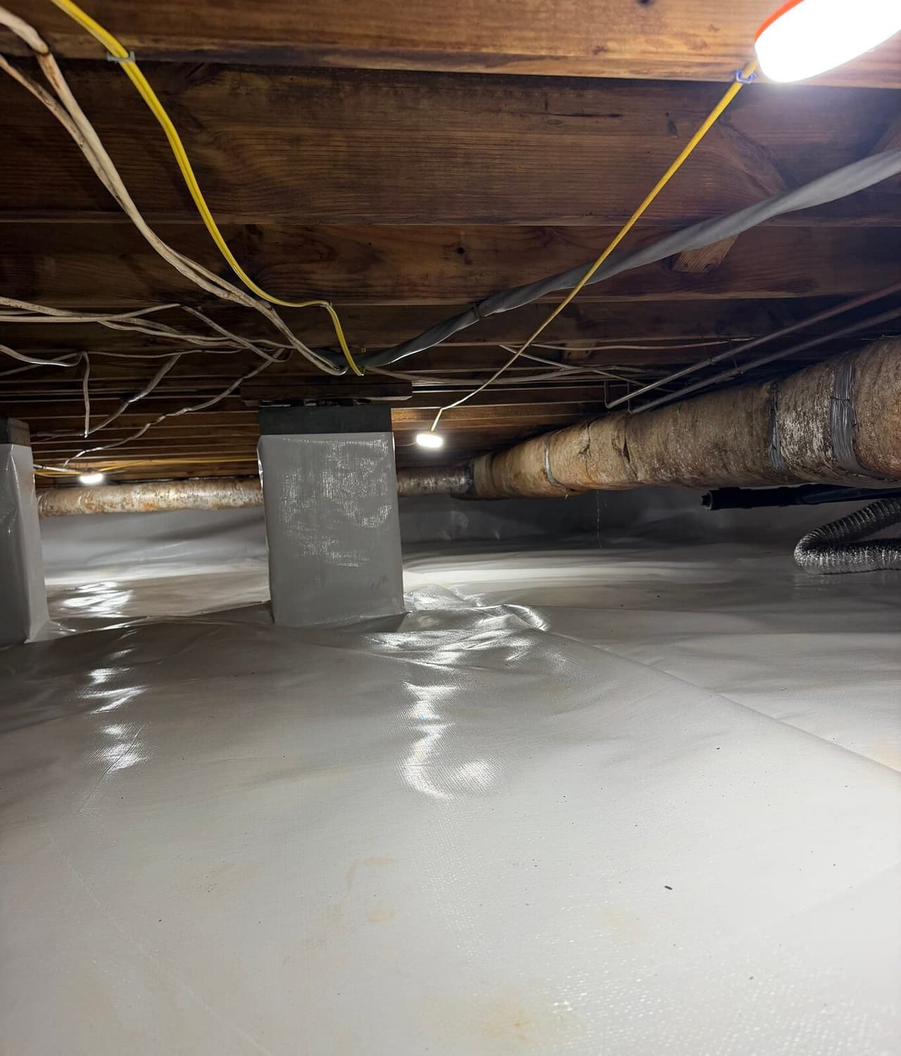 Lined crawl space with wrapped piers and a work light under the floor joists.