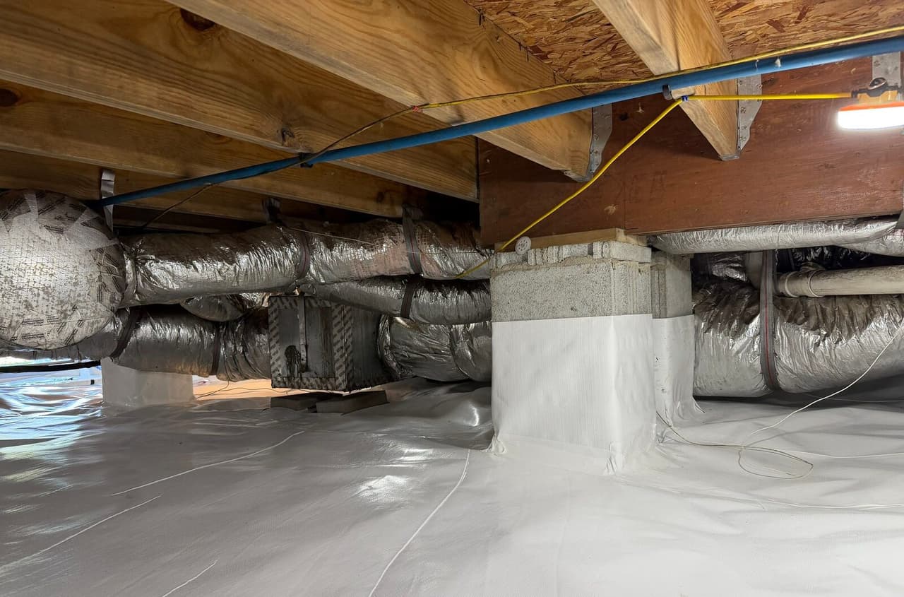 Crawl space with block piers wrapped in white liner and white floor liner beneath silver ducts.
