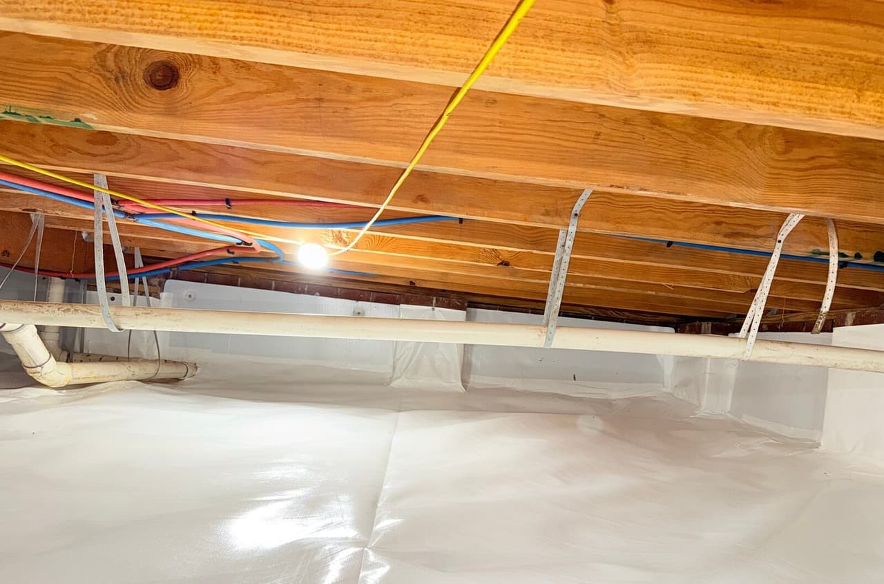 White liner covering crawl space floor and walls below a drain pipe hung from wood joists.
