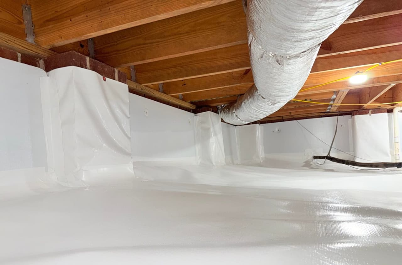 Crawl space with white liner on walls, piers and floor below a silver duct and wood joists.