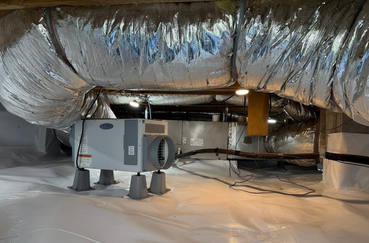 Crawl space dehumidifier on stands over white floor liner beneath insulated ducts and an air handler.