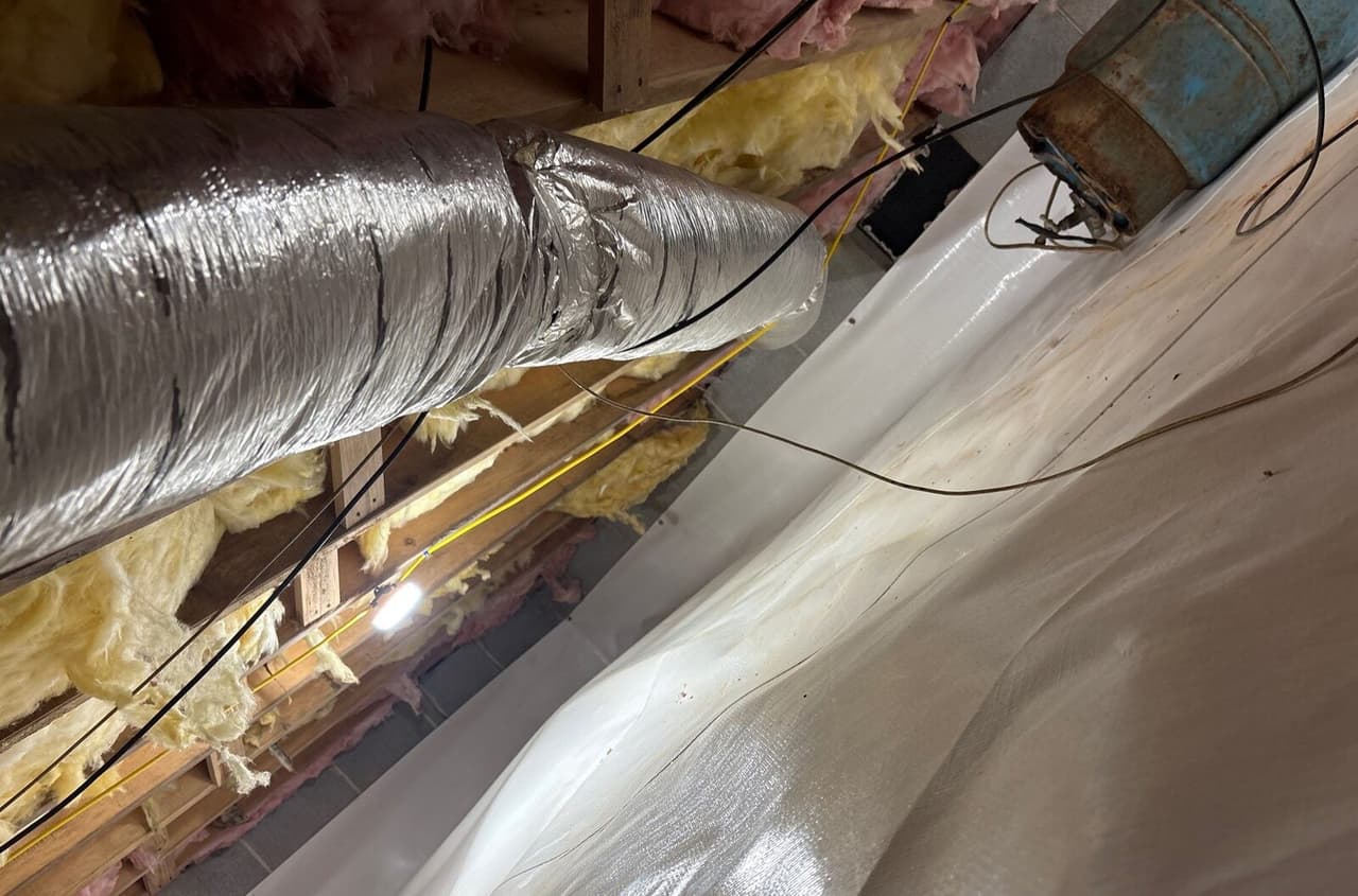 Lined crawl space wall and floor beside insulated ductwork and an old metal tank.