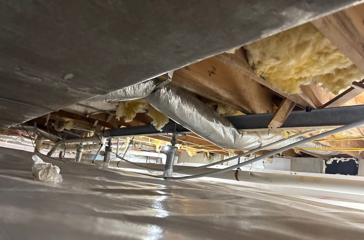 Lined crawl space with a row of steel support posts under a beam, ductwork and pipes overhead.