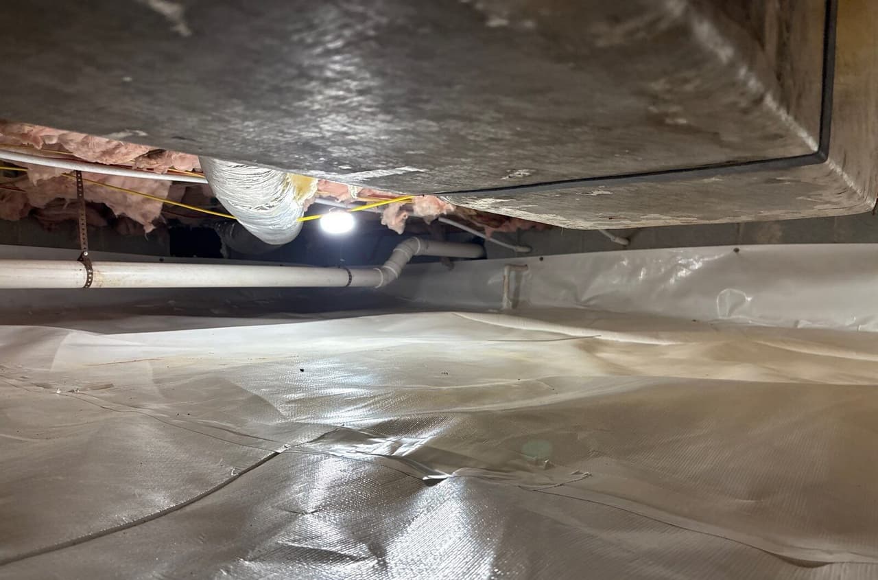 Crawl space with white liner on the floor and walls under a large duct and plumbing pipes.