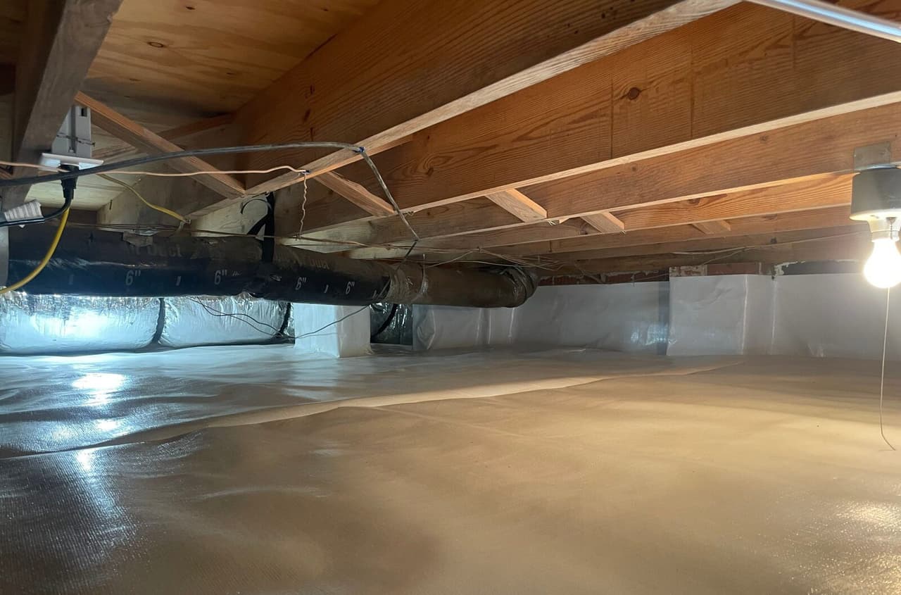 Crawl space with white liner covering the floor and walls under wood joists, ductwork and a hanging bulb.
