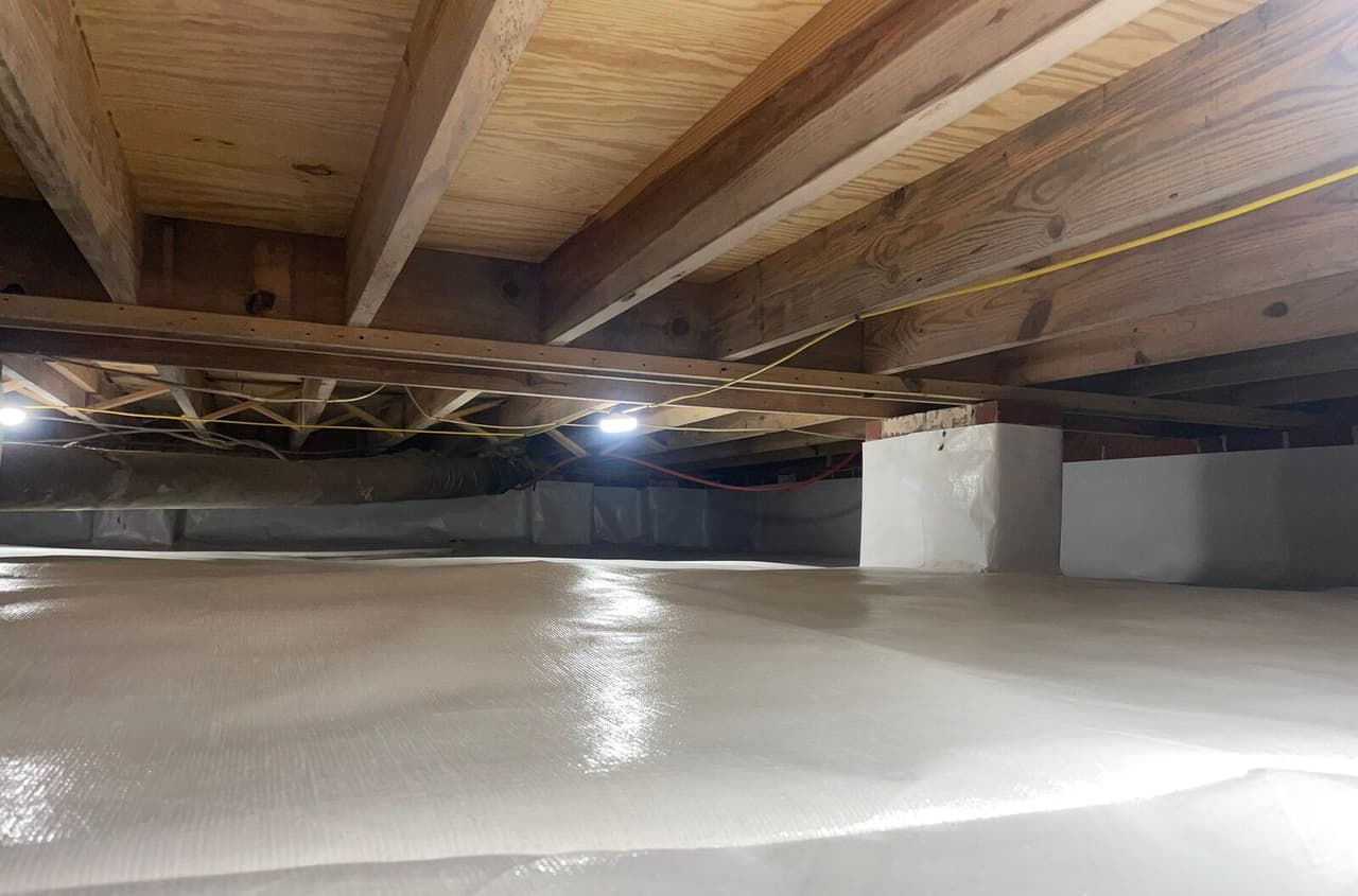 Lined crawl space floor and walls with a wrapped pier under wood joists and lights in the distance.