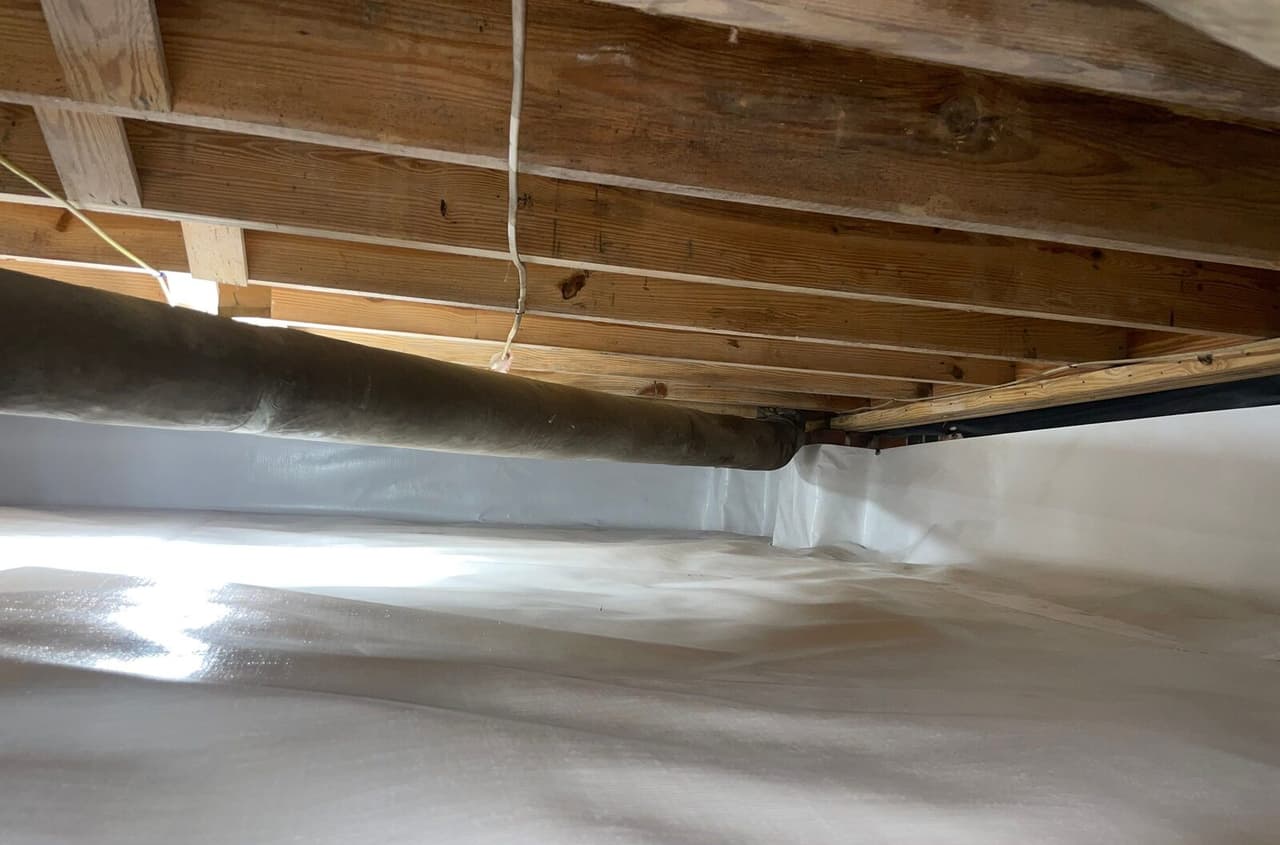 Duct running under joists above a crawl space with liner on the floor and walls.