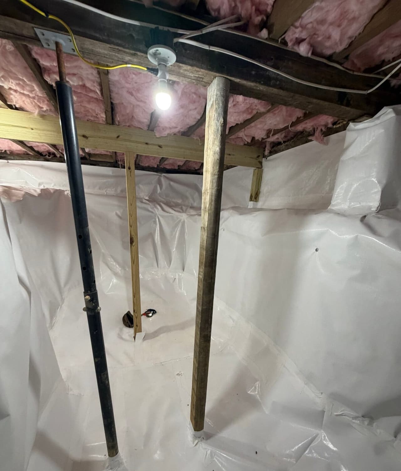 White liner on crawl space walls and floor around a metal post, wood posts and a new beam under a light.