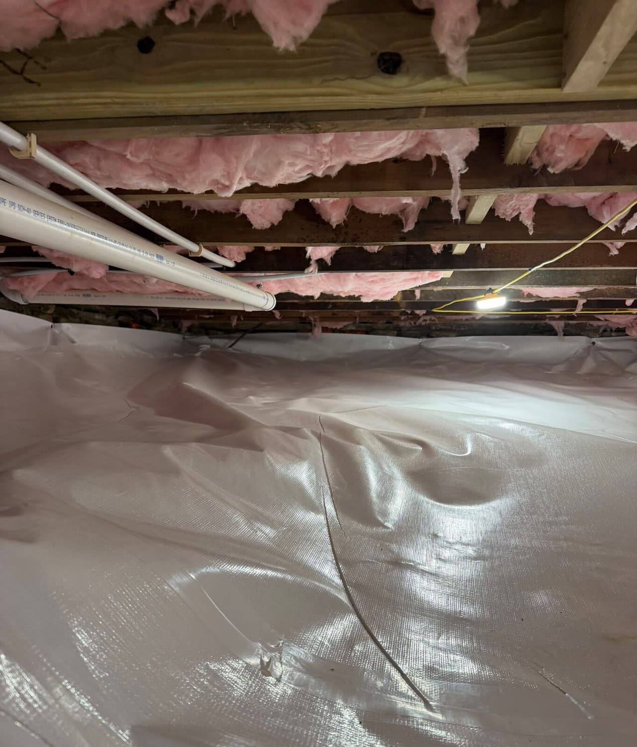 Crawl space floor covered in white liner with a taped seam below plumbing pipes and insulated joists.