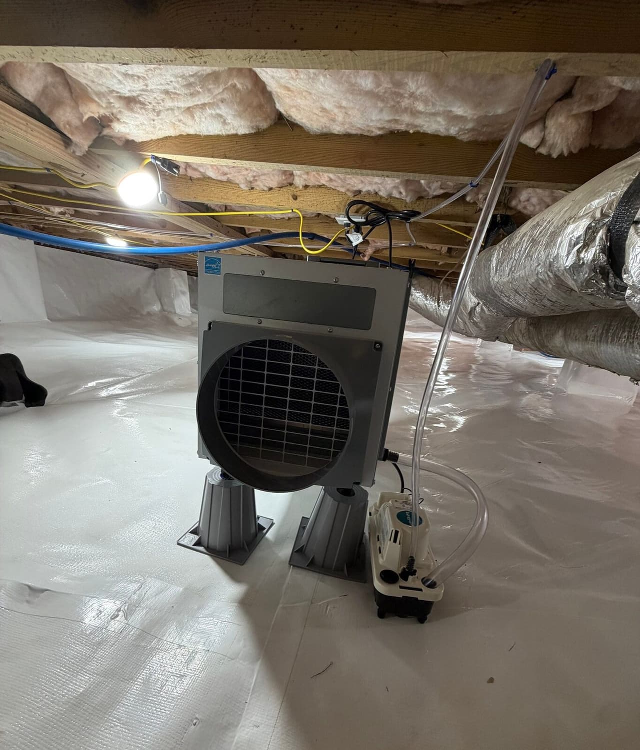 Crawl space dehumidifier on stands with a condensate pump, set on white liner under insulated floor joists.