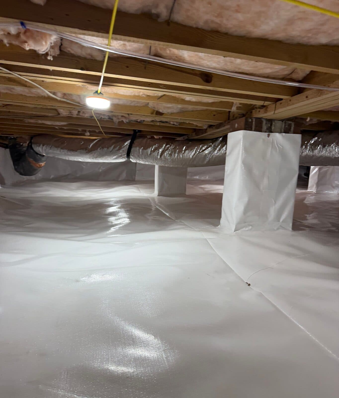 White lined crawl space with wrapped support piers and a silver duct running under the joists.