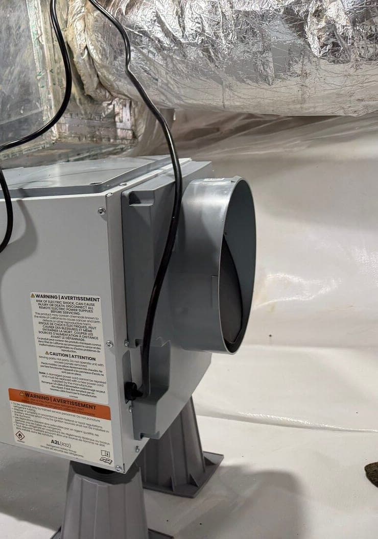 Dehumidifier on raised stands in a lined crawl space with ducts and water tanks behind it.