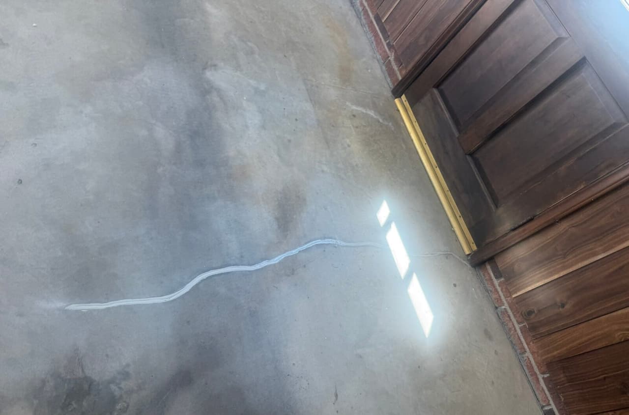 Sealed crack in a concrete floor running toward a wooden door with sunlight on the floor.
