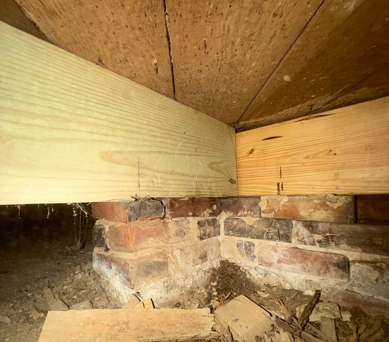 New treated lumber beams resting on an old brick pier in a crawl space with wood scraps below.