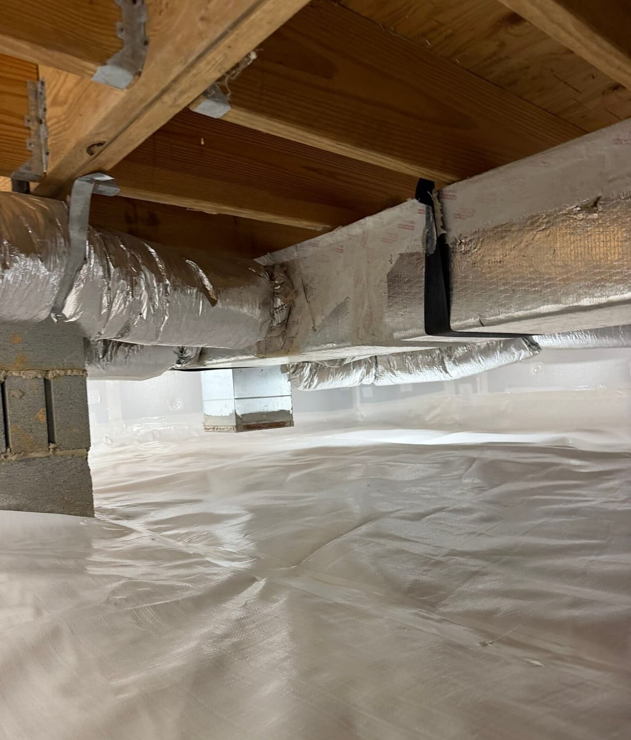 Insulated ducts under floor joists above a crawl space covered in white liner.