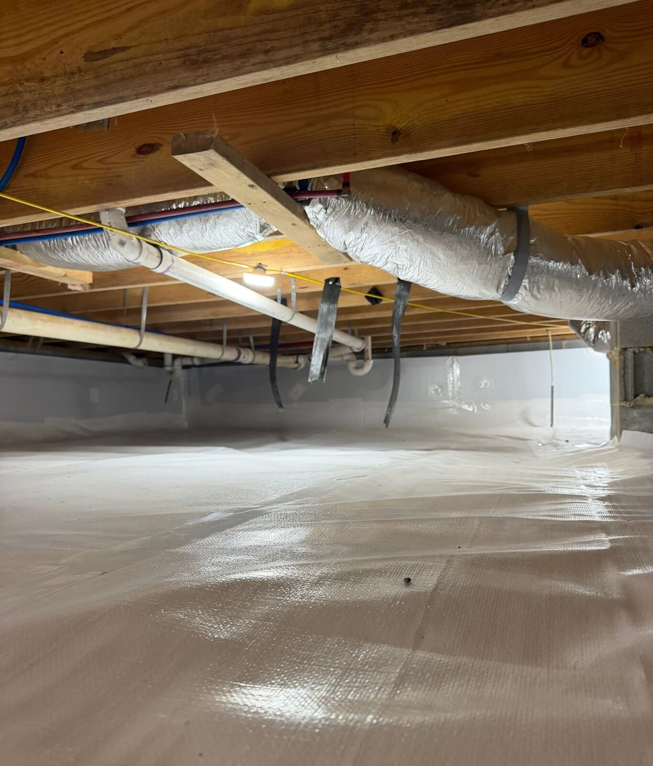 Wide crawl space view with white liner on floor and walls under ducts and plumbing.