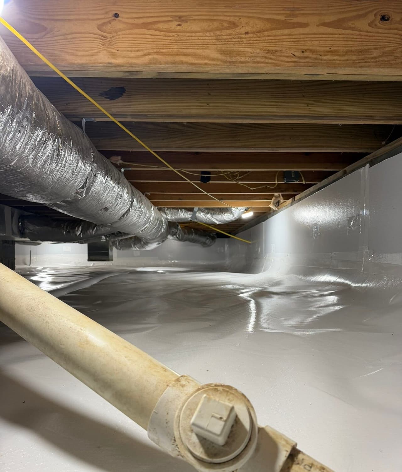 Crawl space with white liner covering the floor and walls beneath ducts and a drain pipe.
