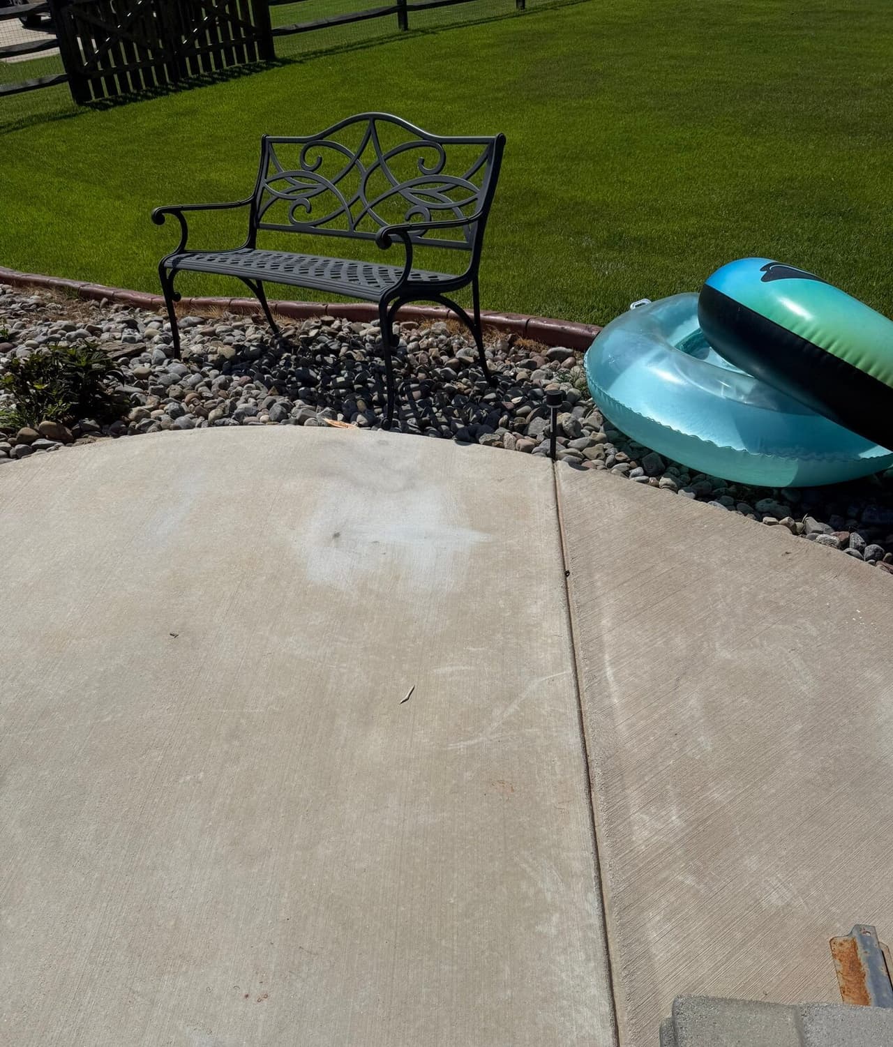Pool deck slabs with a patched injection spot, a metal bench, and pool floats on rock landscaping.
