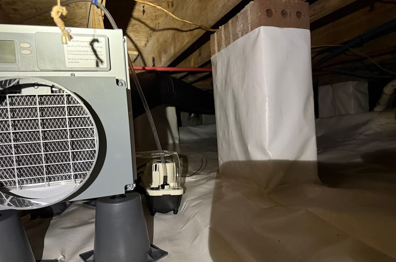 Crawl space dehumidifier on stands with a condensate pump beside a wrapped brick pier and white floor liner.