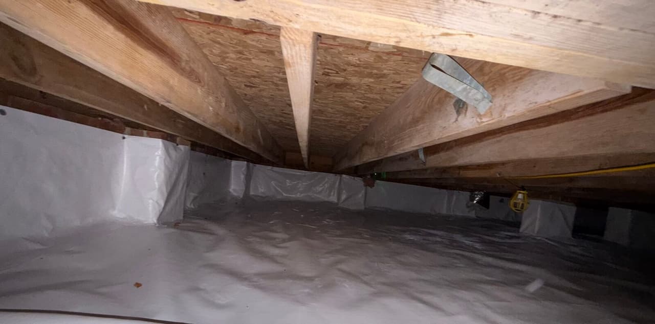 Crawl space with white liner covering the floor and foundation walls beneath wooden floor joists.