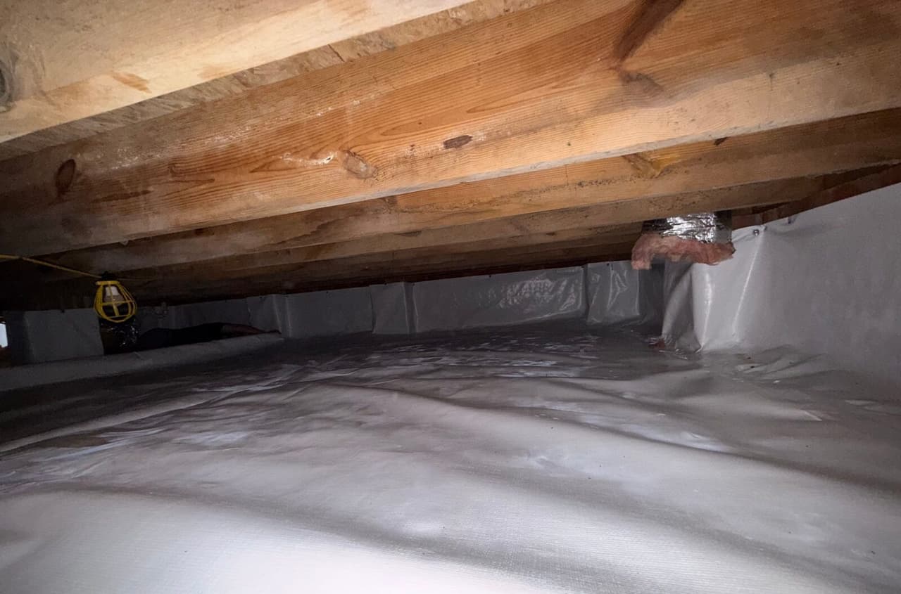 White liner covering a crawl space floor and walls under wooden joists with a work light.