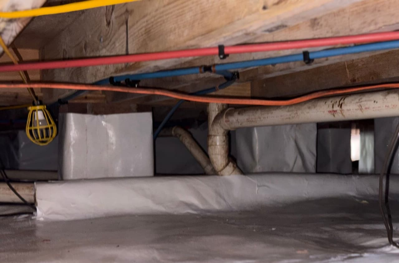 Wrapped piers and white floor liner in a crawl space with pipes and a work light overhead.